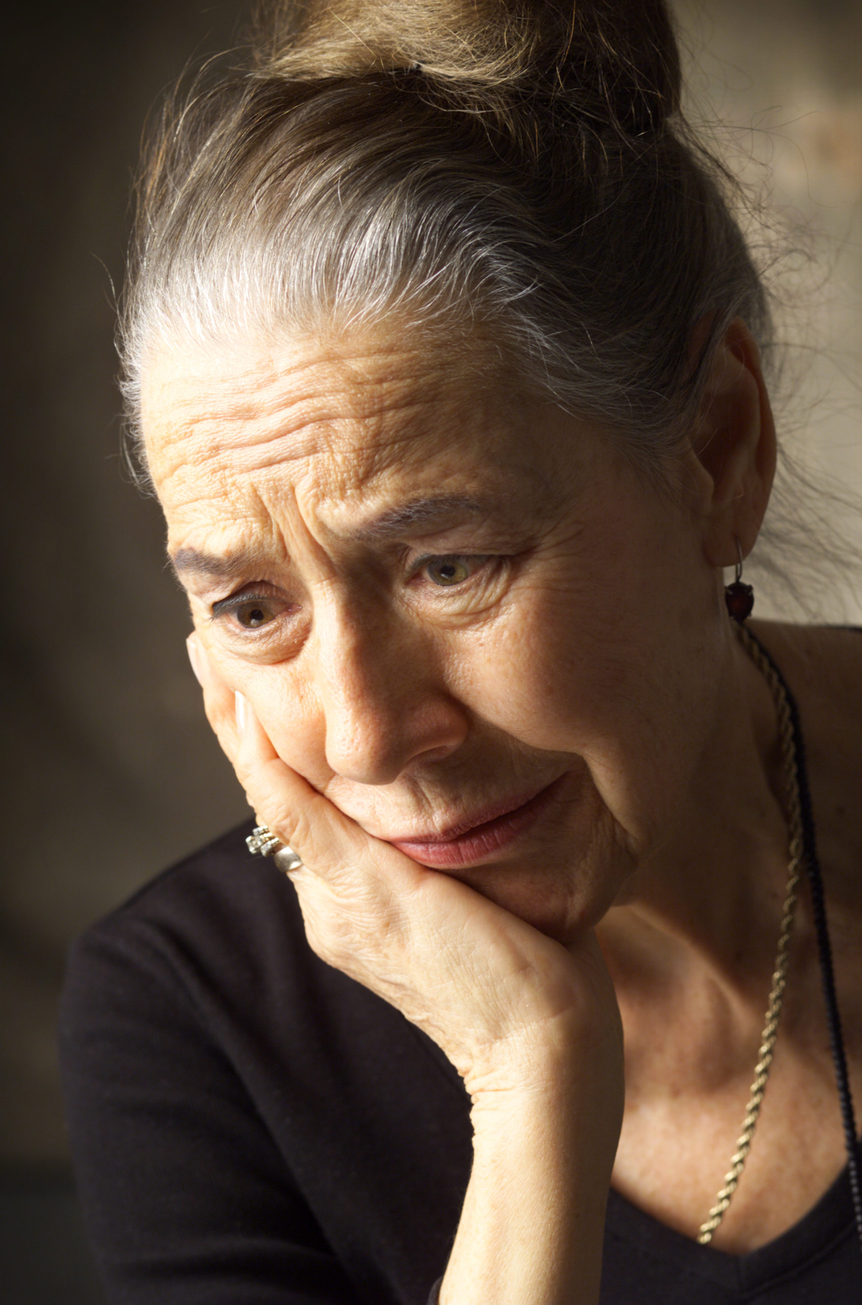an elderly woman with a gray haired bun is wearing a dark dress and in leaning on her hand looking worried